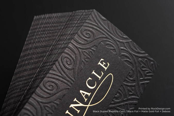 Close-up of a premium black duplex business card with an ornate debossed pattern and elegant matte gold foil logo, showcasing the detailed texture and luxurious finish of the cardstock.