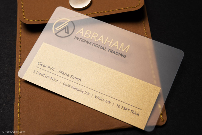Modern gold translucent PVC business card for Abraham International Trading with matte finish, gold metallic ink, and white ink UV print, displayed on brown leather background – RockDesign