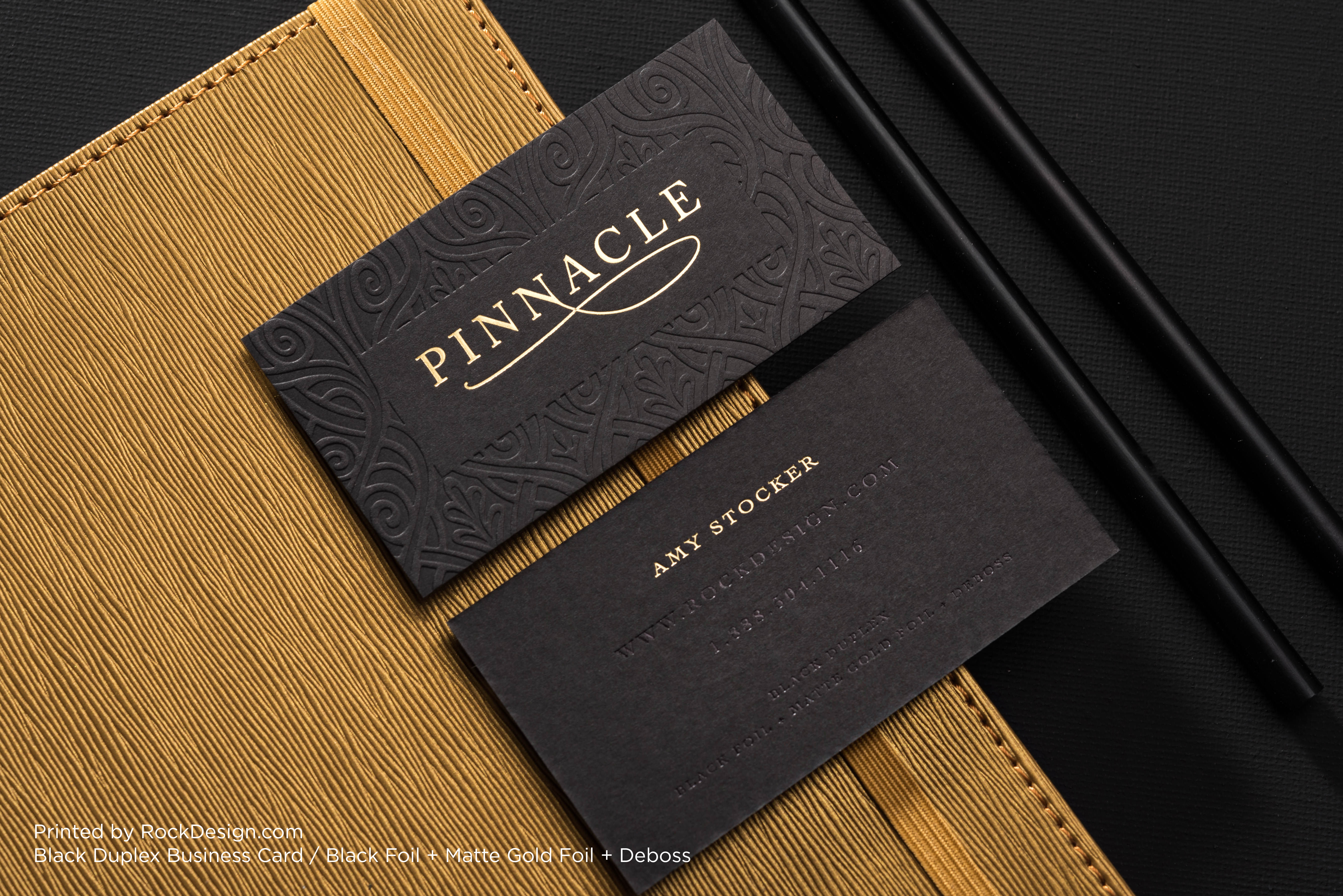Luxury black duplex business card featuring an intricate debossed pattern, black foil lettering, and matte gold foil accents on premium black cardstock, photographed against a textured gold surface.